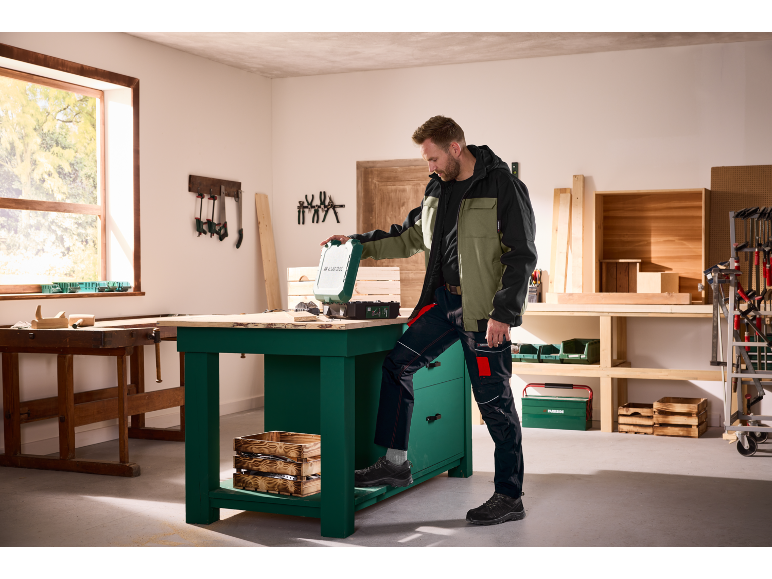 Bărbat în jachetă de lucru și pantaloni cargo într-un atelier, deschizând o cutie verde de scule.