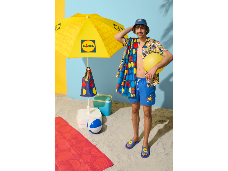 Bărbat pe plajă cu umbrelă, prosop, pantaloni scurți, șlapi și găleată, totul în stil de vară.