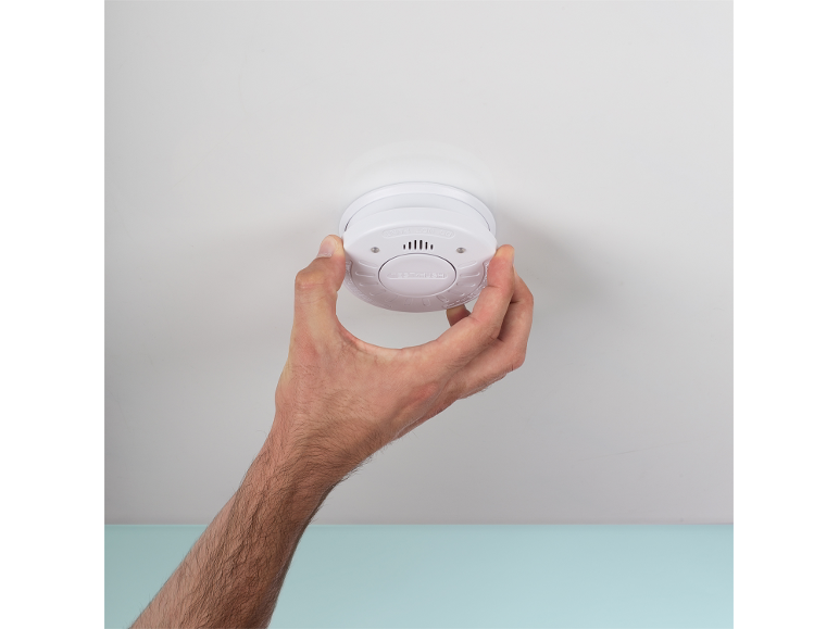 Mână instalând sau testând un detector de fum alb pe tavan.