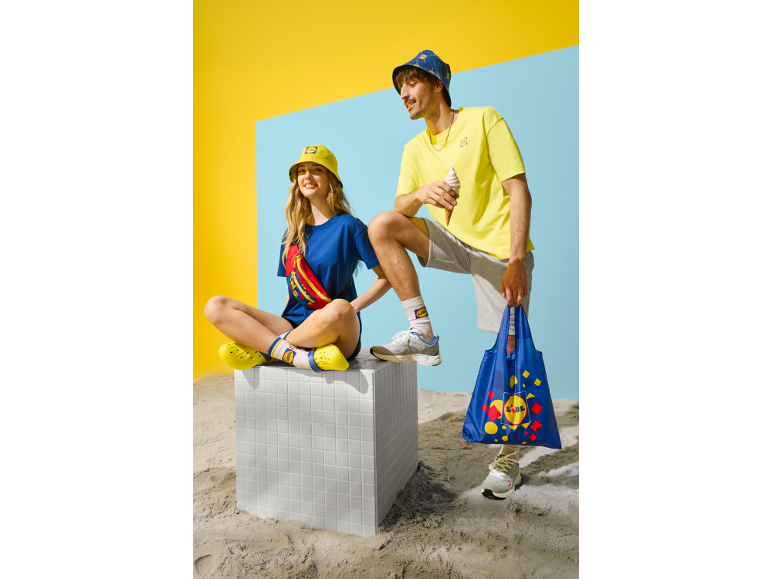 Două persoane în ținute casual de vară, cu pălării tip găleată, tricouri și genți de cumpărături.