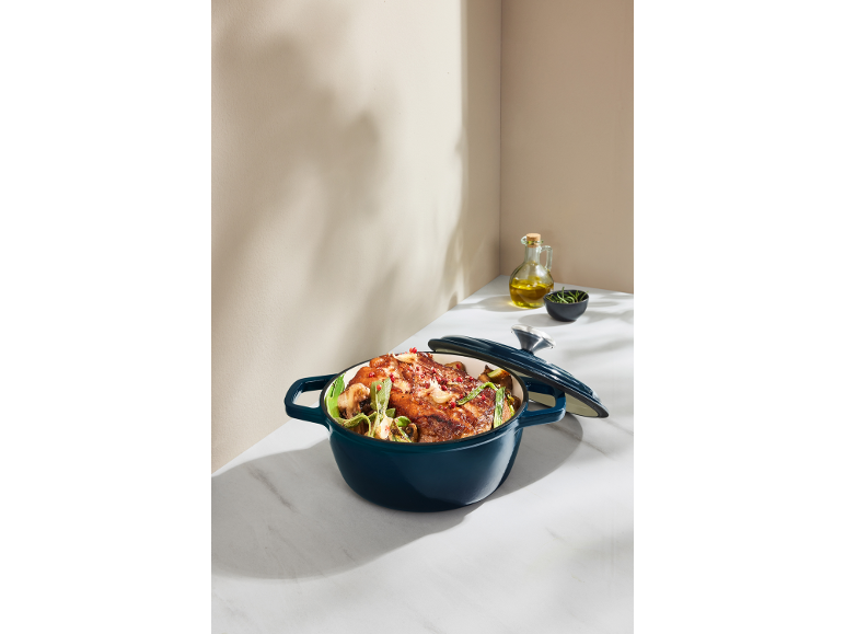 Oală albastră din fontă cu carne și legume, ulei și ierburi pe o suprafață de marmură.