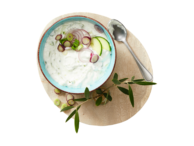 Tzatziki cu castraveți, ceapă roșie și ceapă verde într-un bol, cu o lingură și o ramură de măslin.