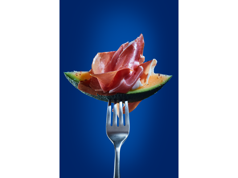 Pepene galben cu prosciutto pe o furculiță, pe fundal albastru.