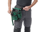 Bărbat în pantaloni de lucru cu o centură verde pentru scule, ținând cuie și un ciocan.