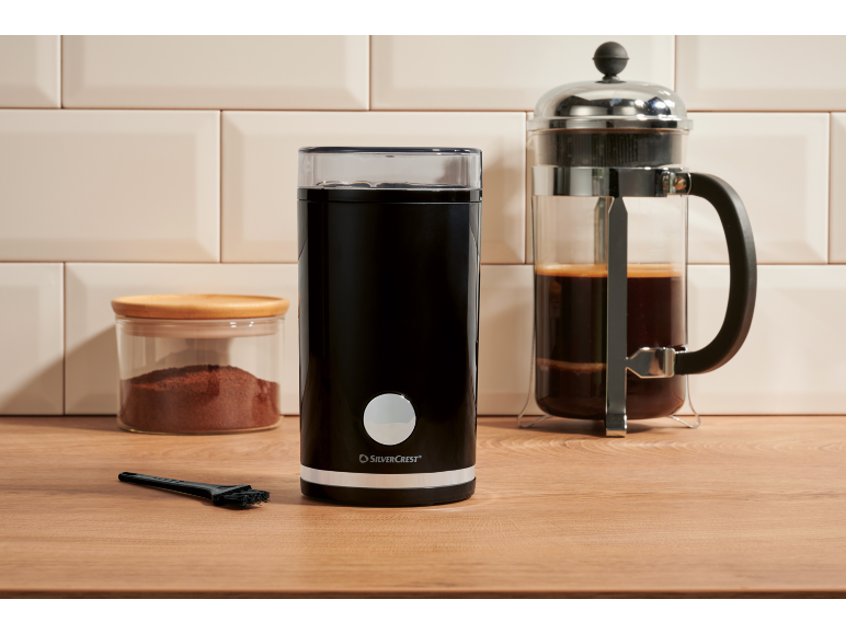 Râșniță de cafea neagră, borcan cu cafea măcinată și presă franceză pe un blat de lemn.