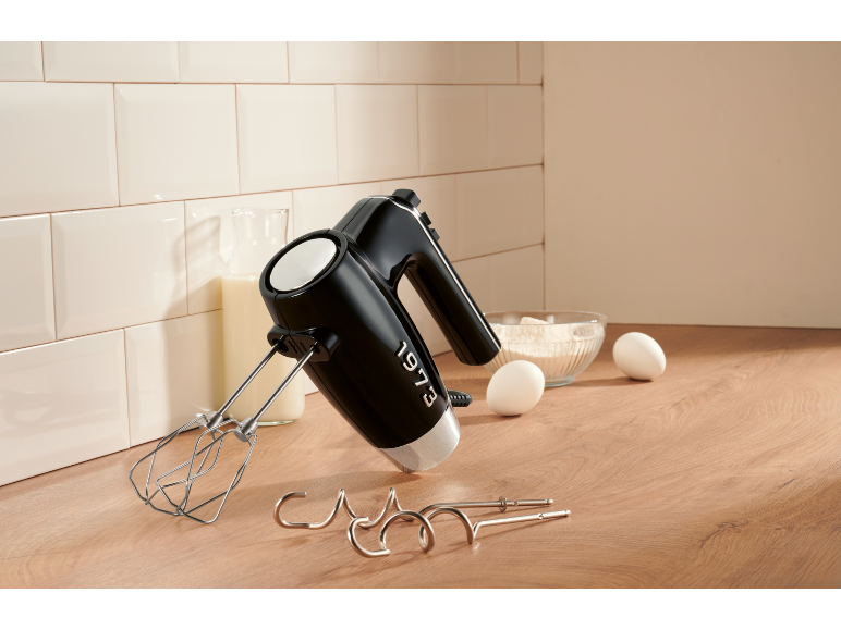 Mixer de mână negru cu accesorii, lapte, făină și ouă pe un blat de lemn.