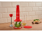 Blender de mână roșu cu accesorii, gata să facă pesto pe un tocător de lemn.