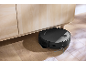 Aspirator robot negru curățând sub un dulap din lemn deschis.
