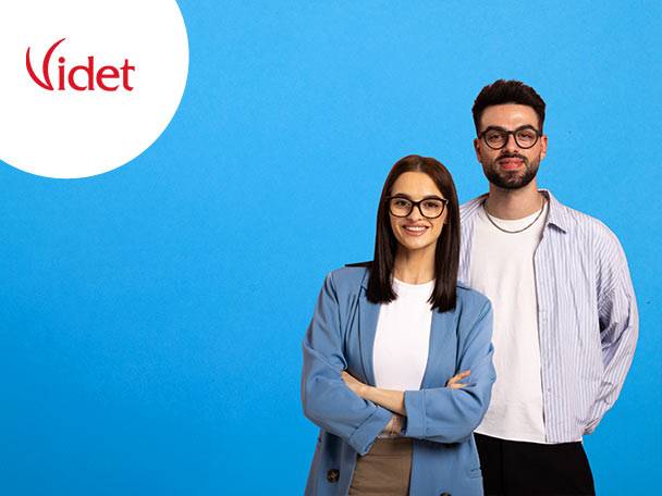 Doi tineri zâmbitori, un bărbat și o femeie, poartă ochelari și stau pe un fundal albastru.