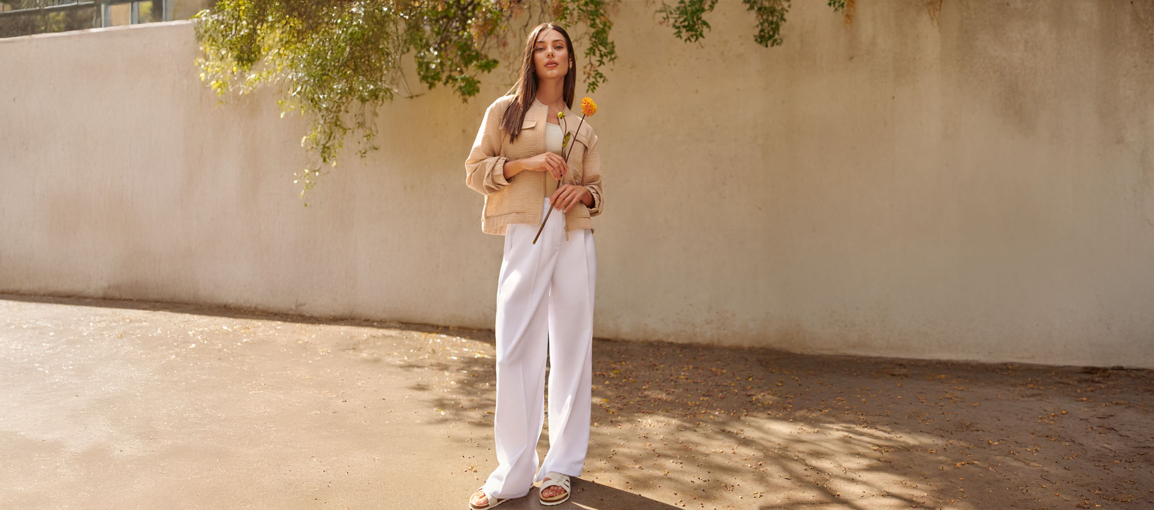 Femeie în jachetă bej, pantaloni albi și sandale, ținând o floare galbenă.