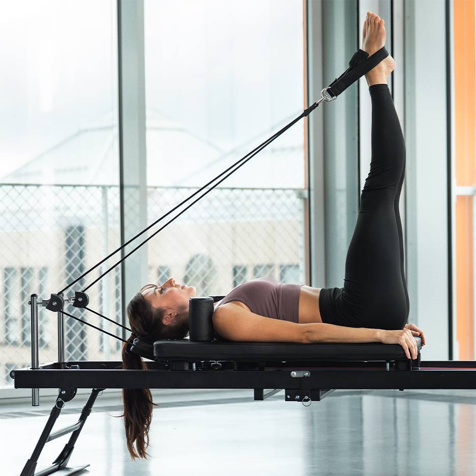 Femeie făcând un exercițiu pentru picioare pe un aparat Pilates reformer.