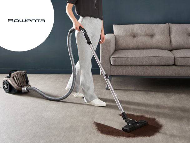 Aspirator cu recipient, curățând o pată de murdărie de pe covor, lângă o canapea.
