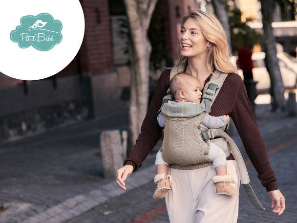 Mamă zâmbitoare purtând un bebeluș într-un marsupiu, plimbându-se pe stradă. Logo: Petit Bébé.