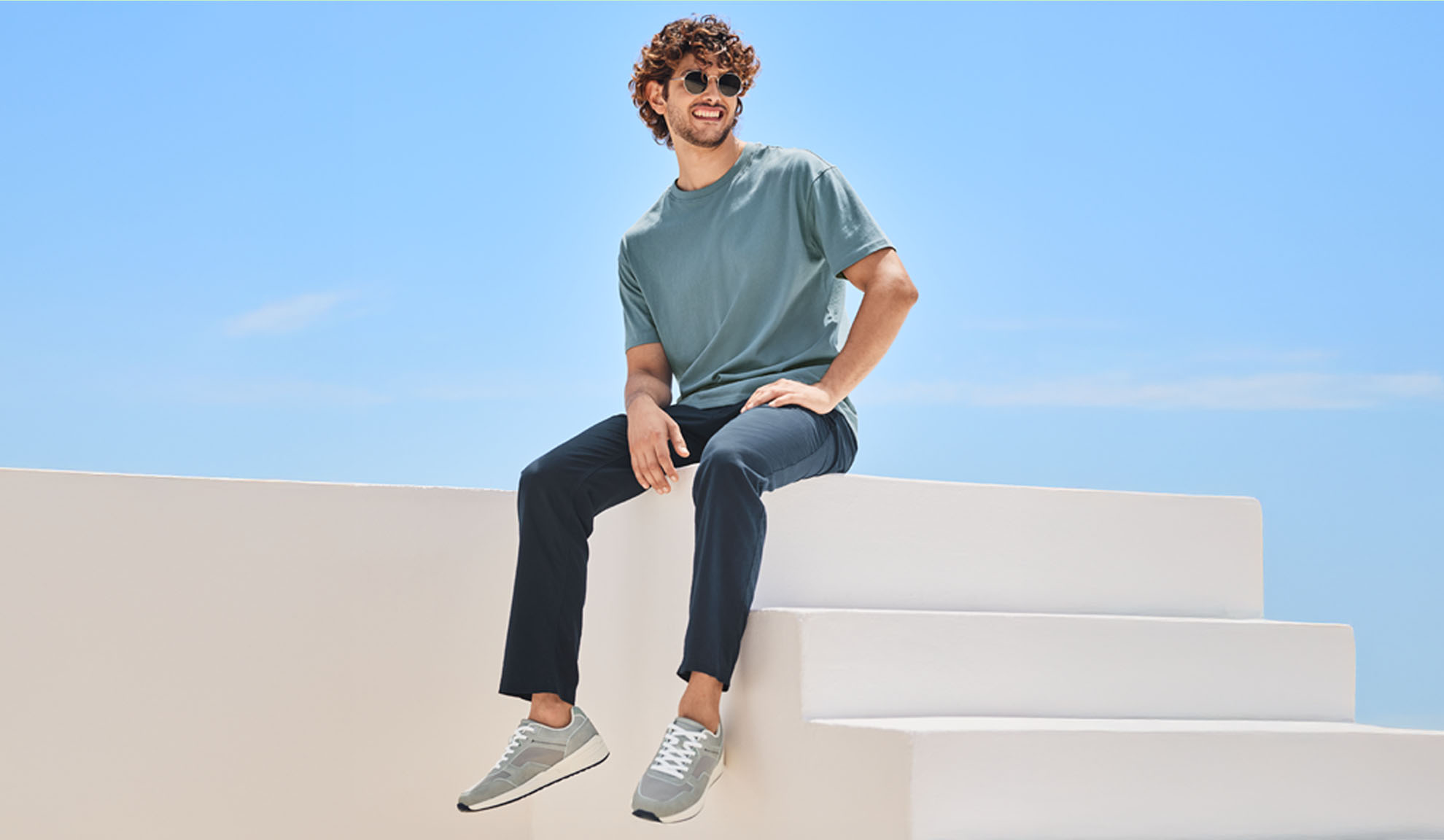 Un bărbat relaxat îmbrăcat într-un tricou și pantaloni bleumarin, așezat pe o scară albă.