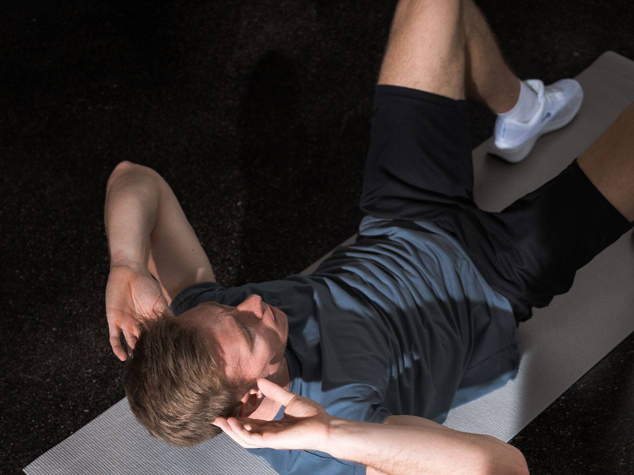 Bărbat în tricou albastru și pantaloni scurți negri face abdomene pe o saltea de yoga.