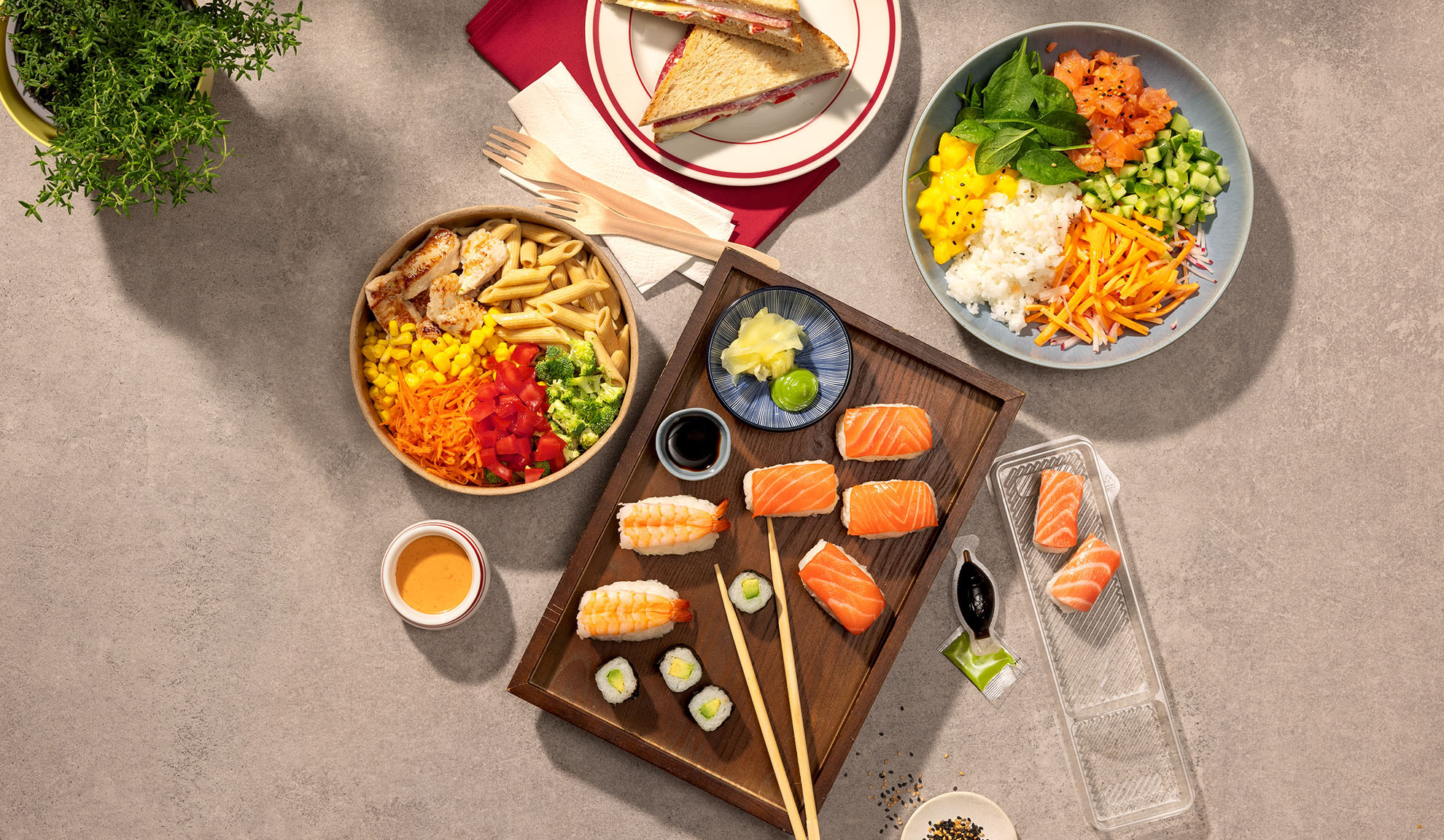 Vedere de sus a diverselor mese la pachet: sushi, poke bowl, salată de paste și sandvișuri.