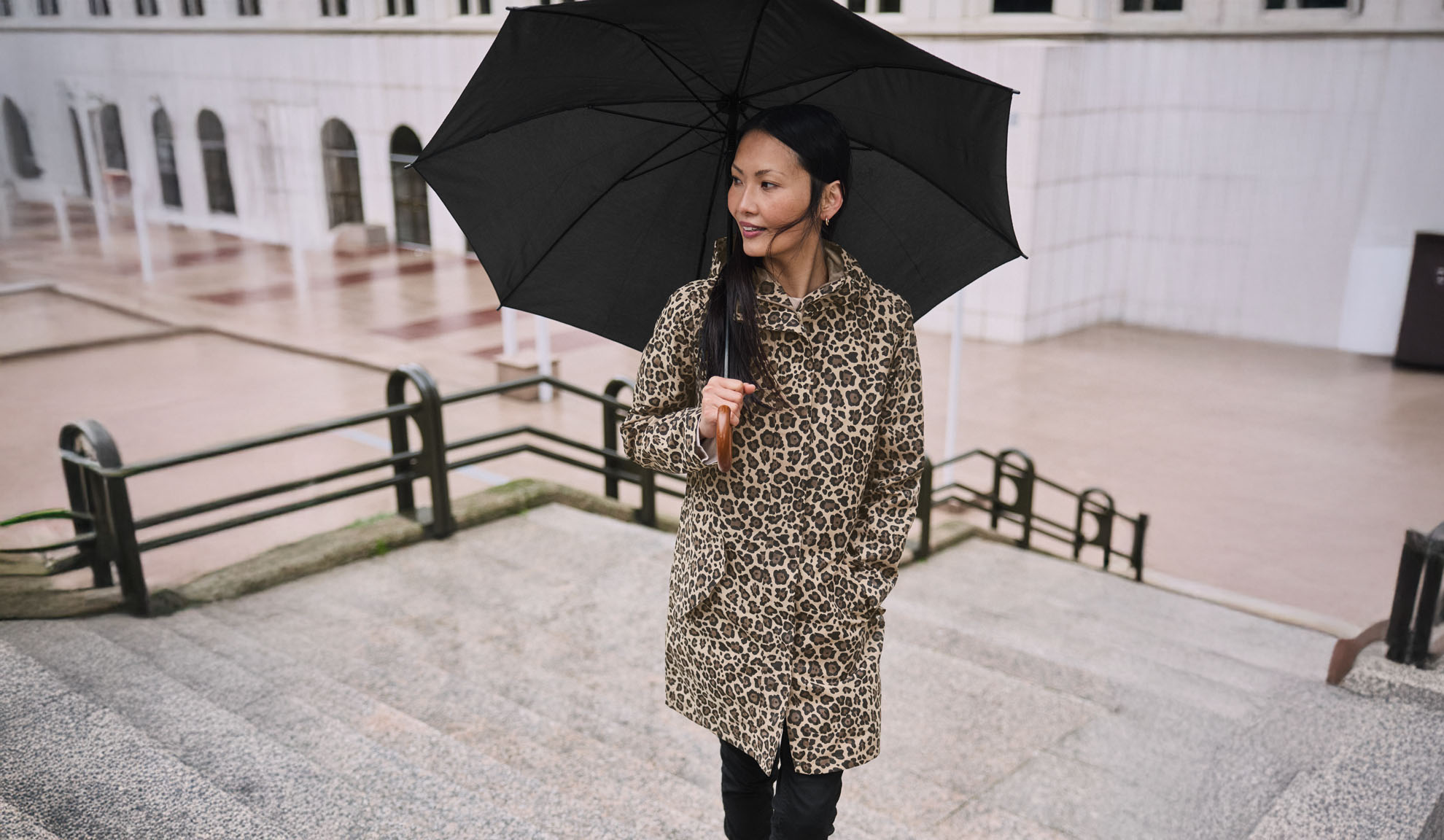 O femeie într-un palton leopard ține o umbrelă neagră pe o scară.