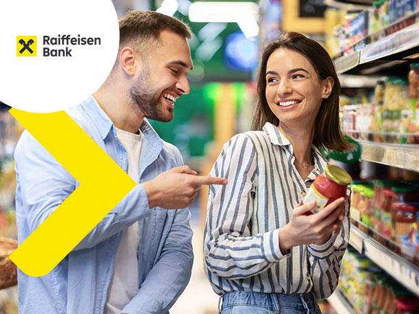 Un cuplu zâmbitor într-un supermarket, bărbatul arătând spre un borcan de sos ținut de femeie.