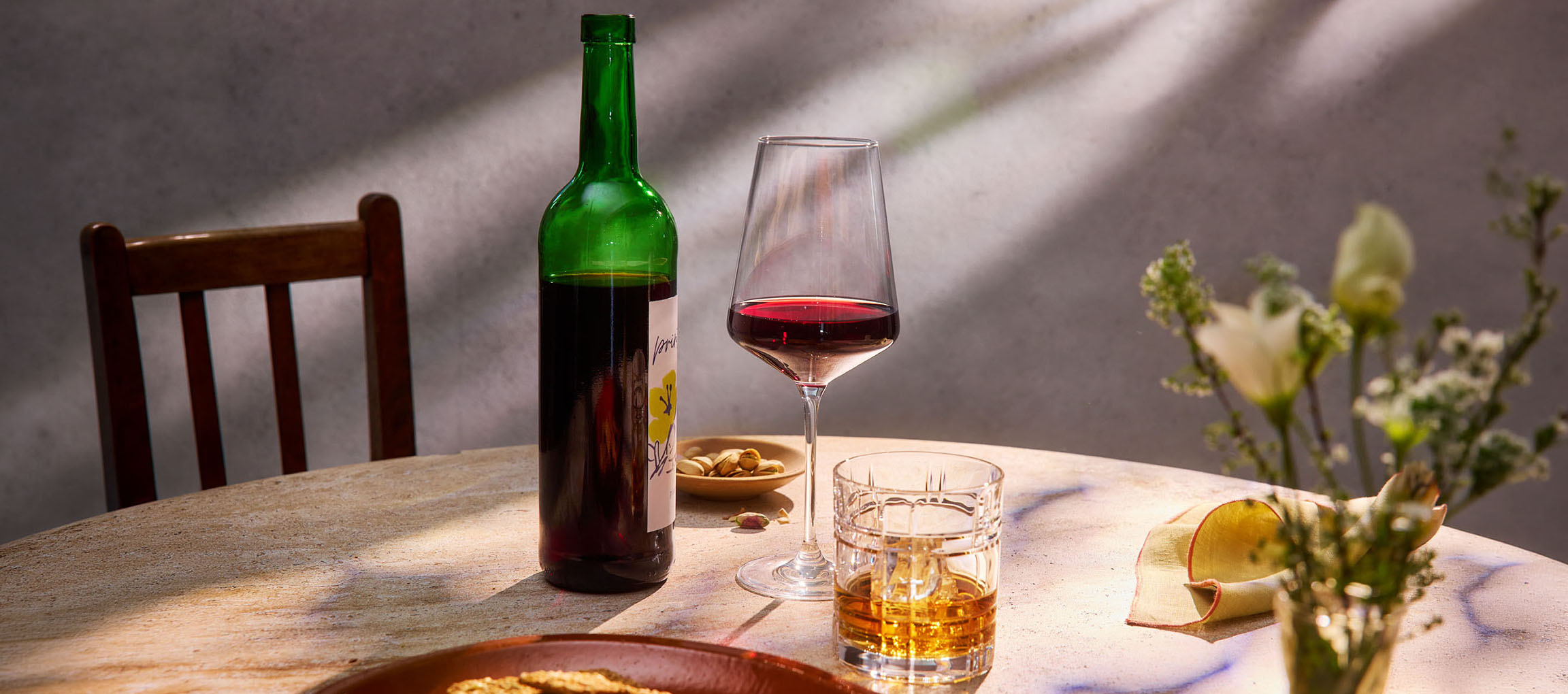 Sticlă de vin roșu, pahar de vin și whisky pe masă, cu scaun și flori în fundal.