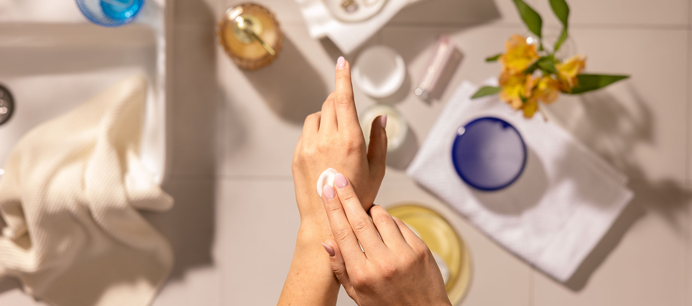 Mâini de femeie aplicând cremă de mâini, înconjurate de cosmetice și prosoape într-o baie.