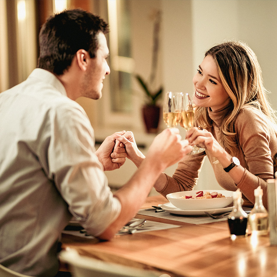 Cuplu la cină romantică, ținându-se de mână și ciocnind pahare cu vin.