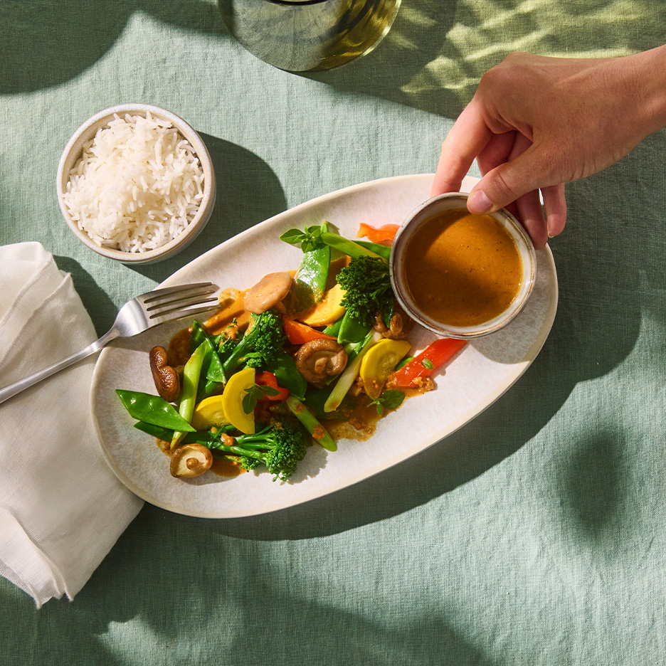 Mâncare asiatică cu legume, ciuperci, orez și sos de arahide pe o masă verde.