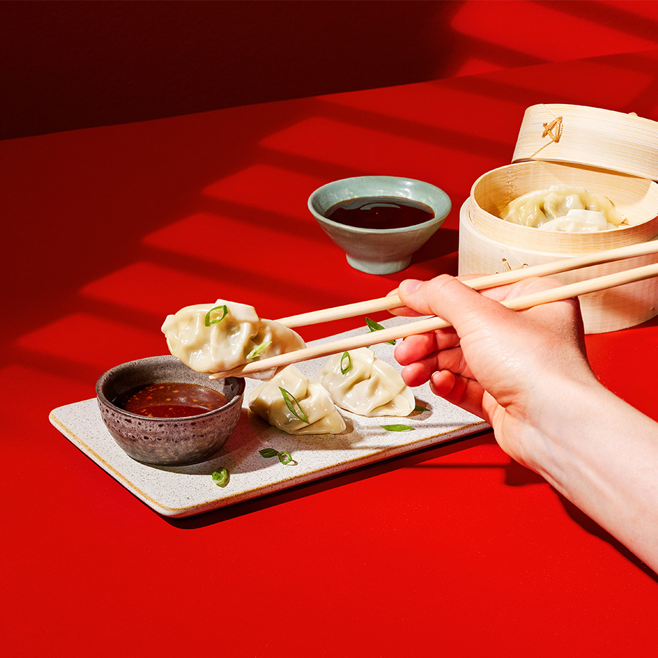 Găluște chinezești cu sos, luate cu bețișoare, pe o masă roșie.