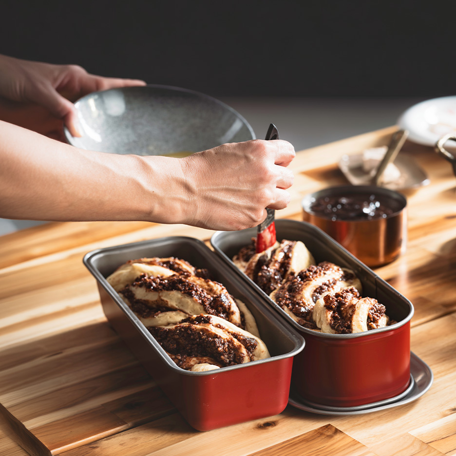 Mână care unge cu pensula două pâini dulci cu ciocolată în tăvi roșii, pe o masă de lemn.