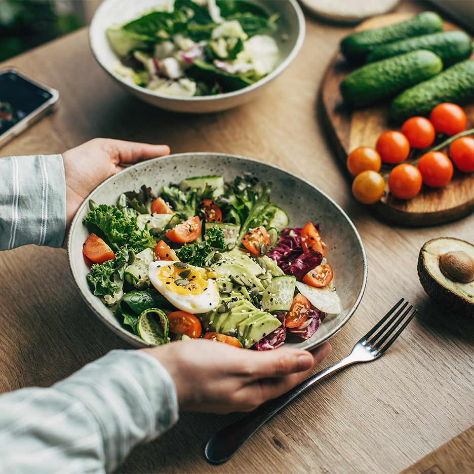 O persoană ține un bol cu salată proaspătă, cu ou, avocado și roșii cherry, pe o masă de lemn.