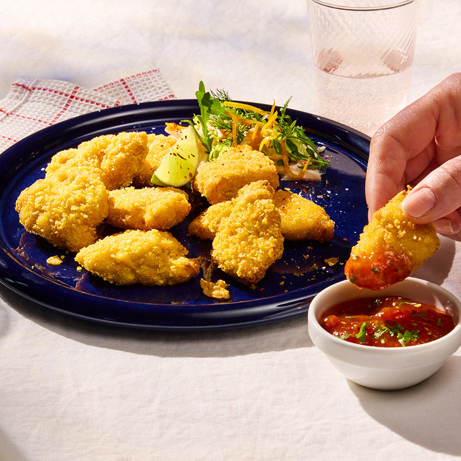 Bucățele de pui pane aurii pe o farfurie albastră, cu salată, lime și sos, o mână înmoaie o bucată în sos.