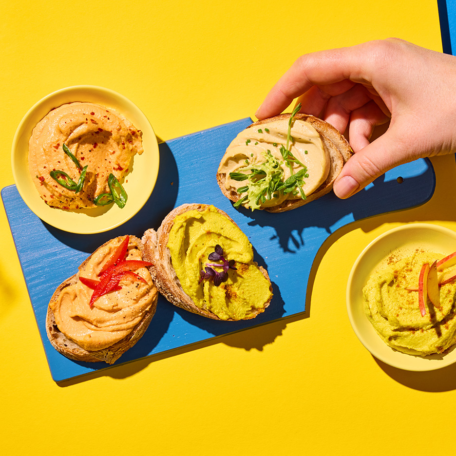 Tartine cu hummus de diferite culori și arome, pe o placă albastră și un fundal galben.
