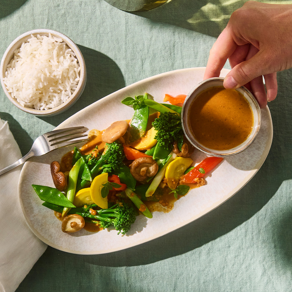 Legume sotate cu sos de arahide și orez, cu o mână care toarnă sos.