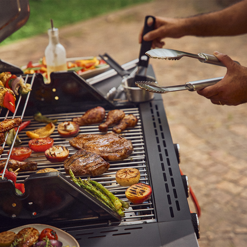 Grătar cu carne, legume și fructe la grătar, cu o persoană care folosește clești.