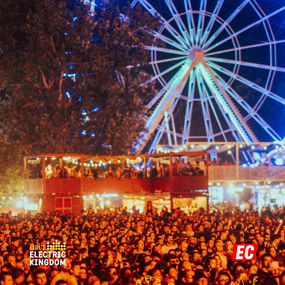 Mulțime de oameni la un festival de muzică, cu o roată mare și o clădire cu două etaje în fundal.