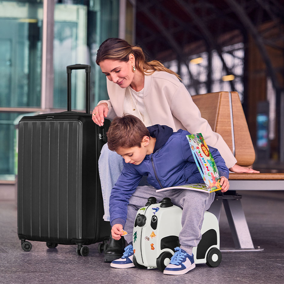 Femeie și copil cu o valiză mare neagră și o valiză-trotineta panda într-un aeroport.