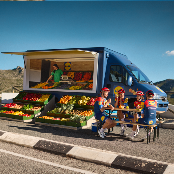 Cicliști se odihnesc lângă un camion Lidl încărcat cu fructe și legume.