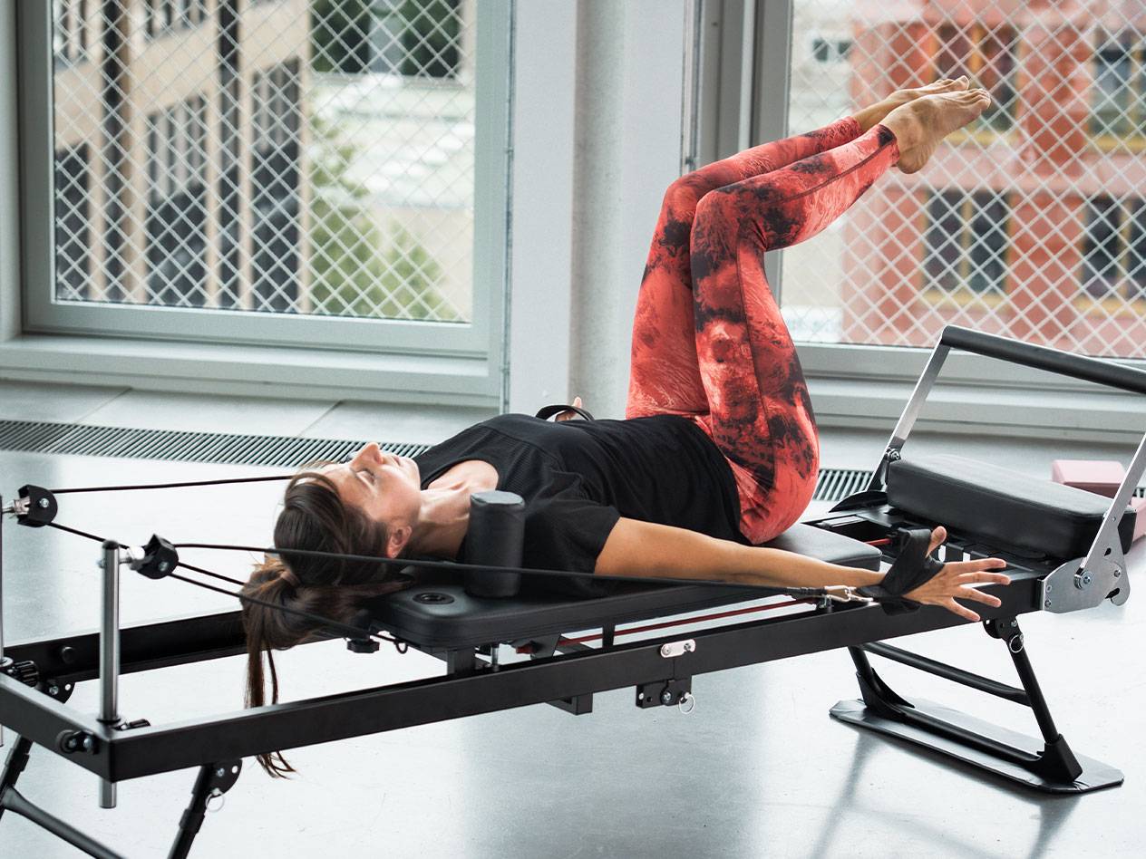 Femeie făcând Pilates pe un aparat reformer, în colanți roșii și negri