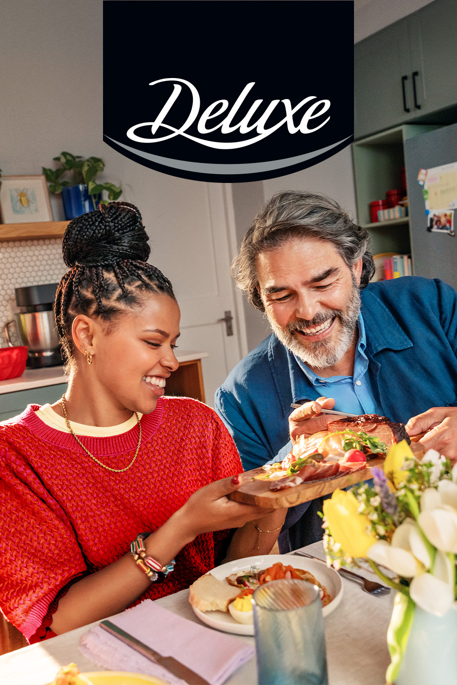 Un cuplu zâmbitor se bucură de o masă cu platouri de carne și legume, cu logo-ul „Deluxe”.