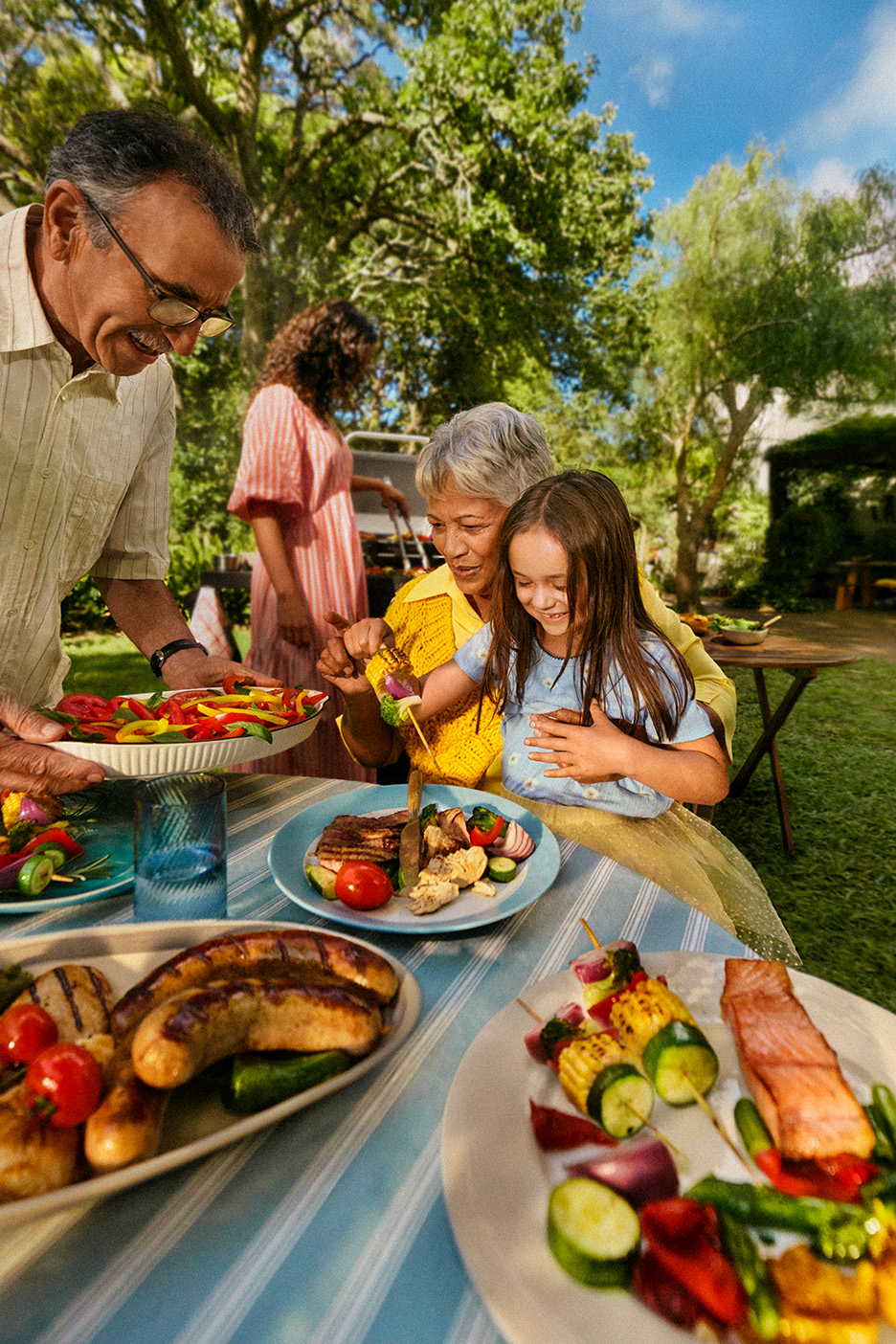 Familie la grătar în grădină, cu cârnați, somon și frigărui de legume.