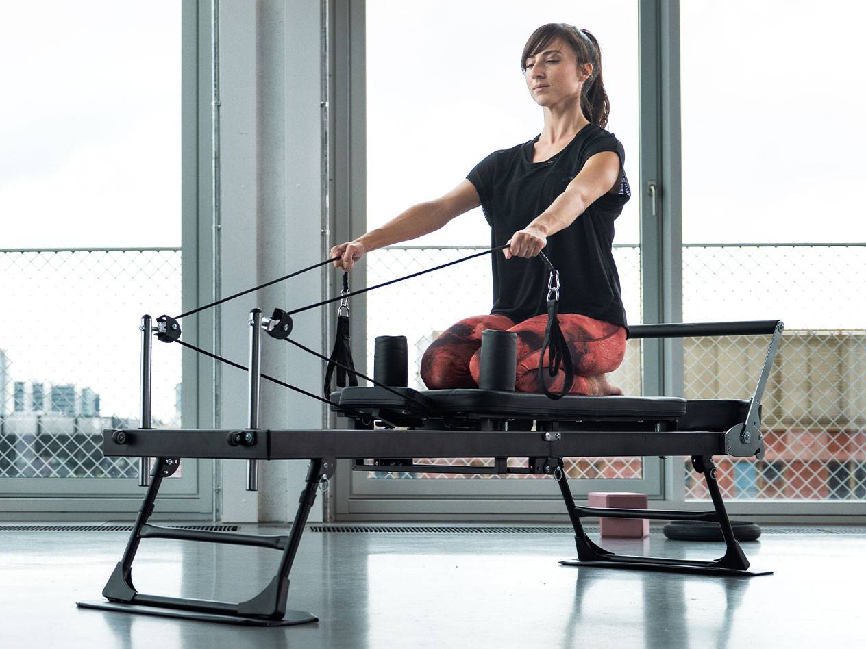 Femeie făcând Pilates pe un aparat reformer, cu fundal de ferestre mari.