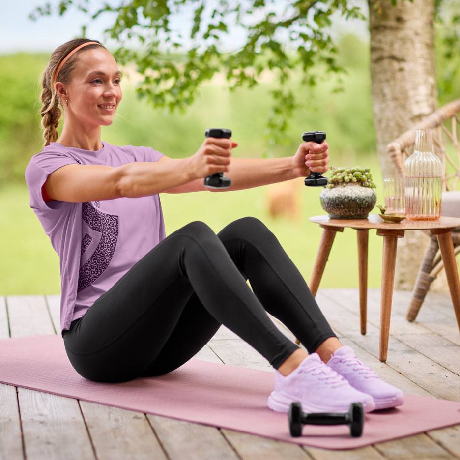 Femeie în tricou mov și colanți negri, făcând exerciții cu gantere pe un covor de yoga.