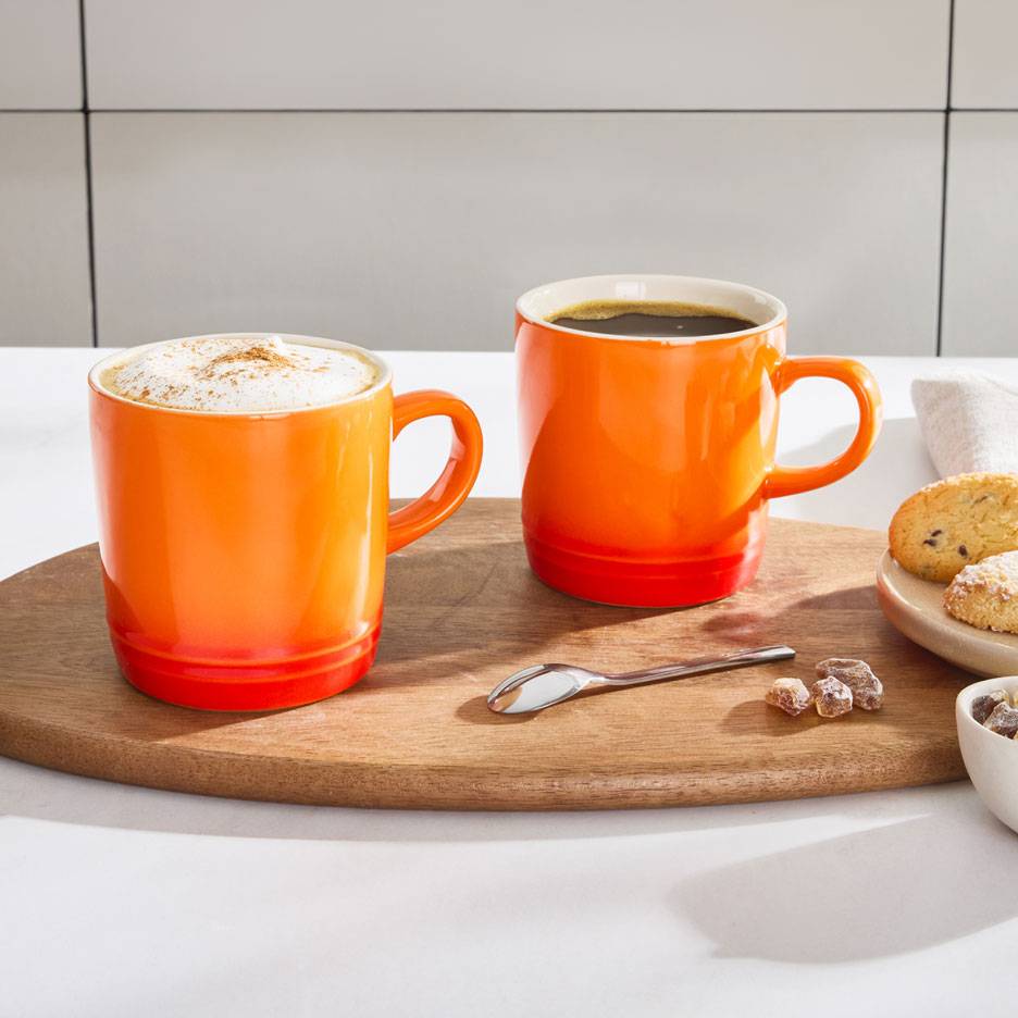 Două căni de cafea portocalii pe o tavă de lemn, una cu cafea spumoasă, cealaltă cu cafea neagră.