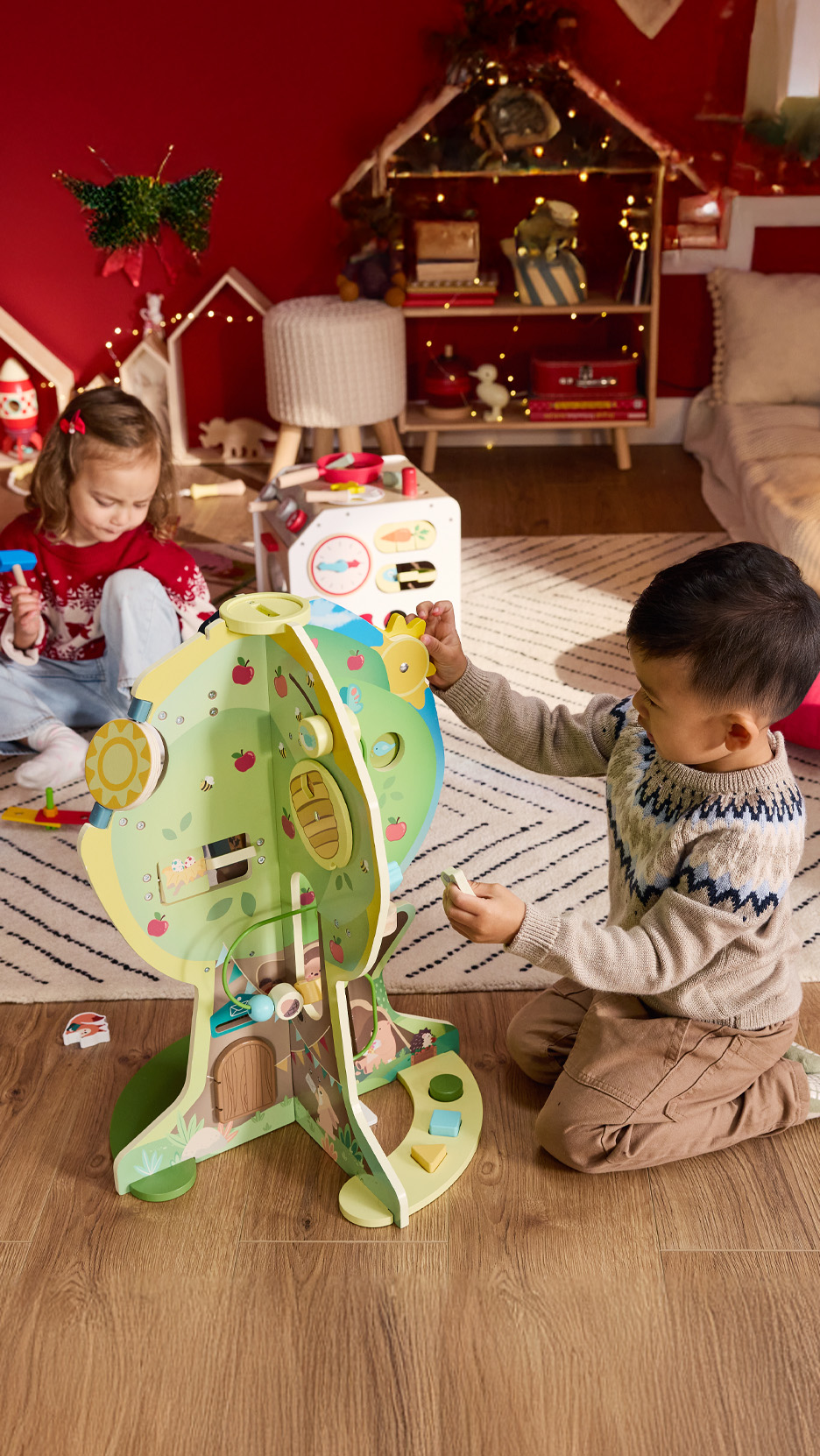 Doi copii se joacă cu o casă de păpuși din lemn în formă de copac și alte jucării.