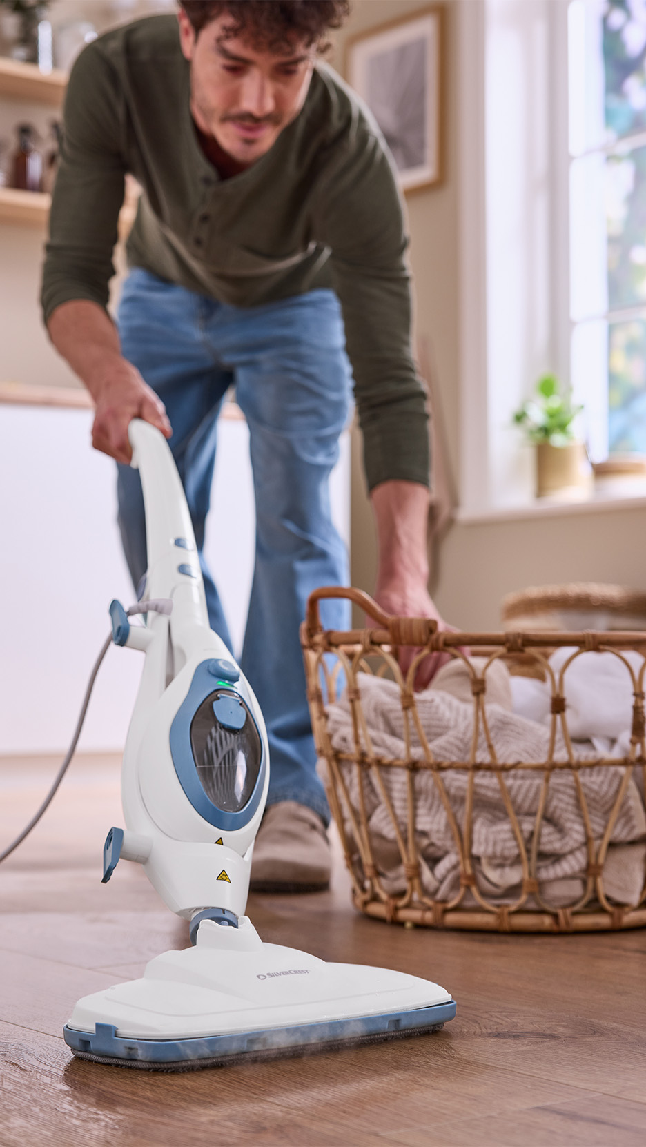Bărbat curățând podeaua cu un mop cu aburi SilverCrest, lângă un coș de rufe.