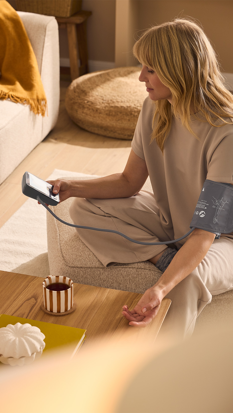 Femeie măsurându-și tensiunea arterială cu un tensiometru, așezată pe o canapea.