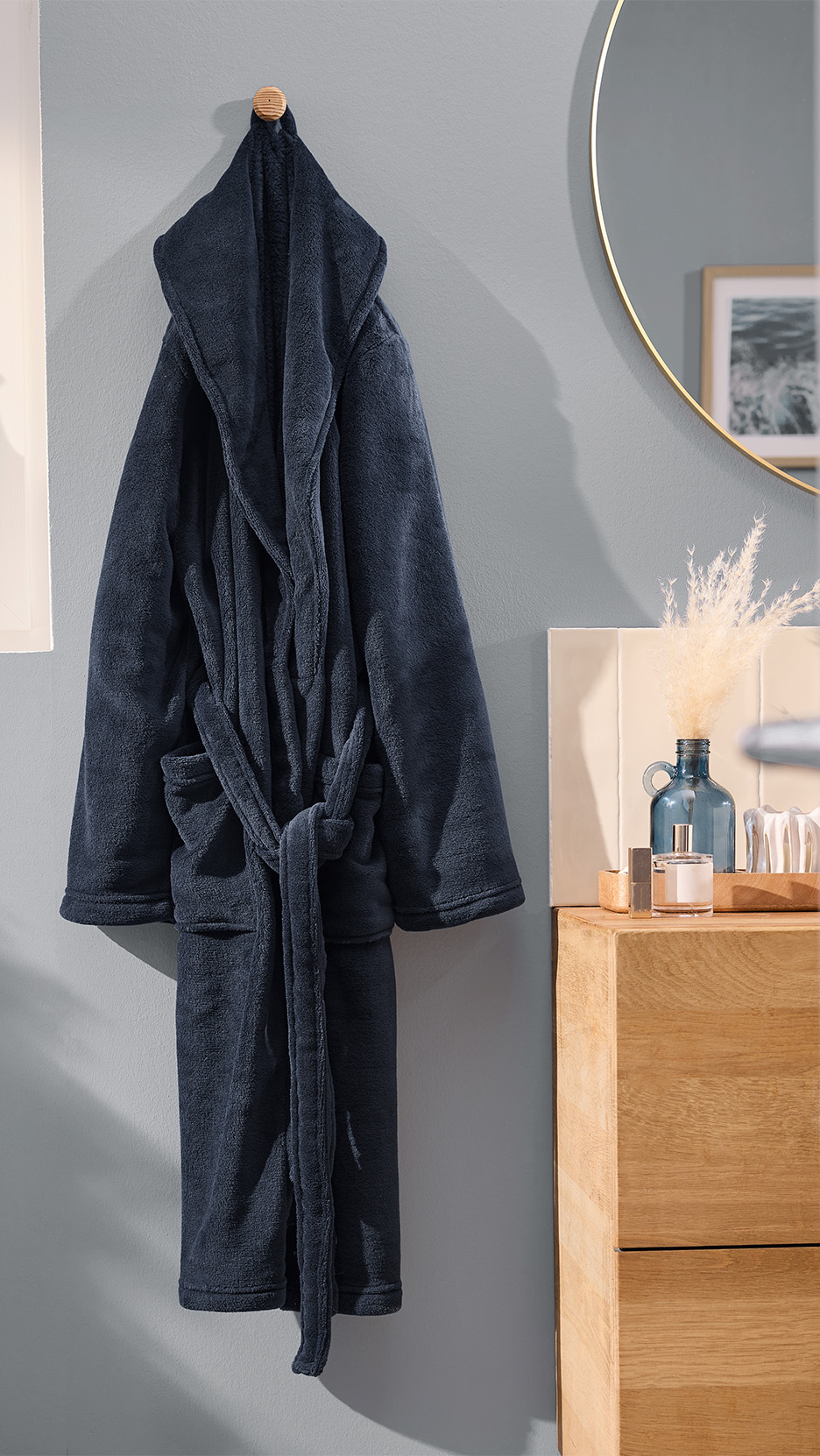 Halat de baie bleumarin agățat pe un perete gri, cu oglindă și dulap din lemn.