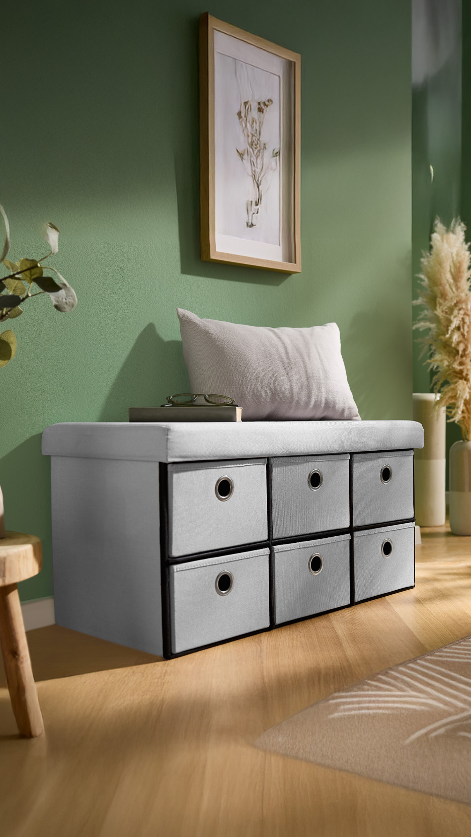 Bancă de depozitare gri cu șase sertare, pernă și decorațiuni pe un perete verde.
