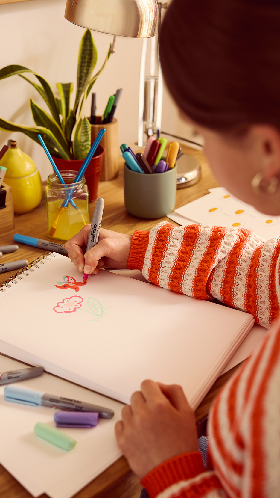 O persoană desenează cu markere colorate pe o carte de schițe, pe un birou din lemn.