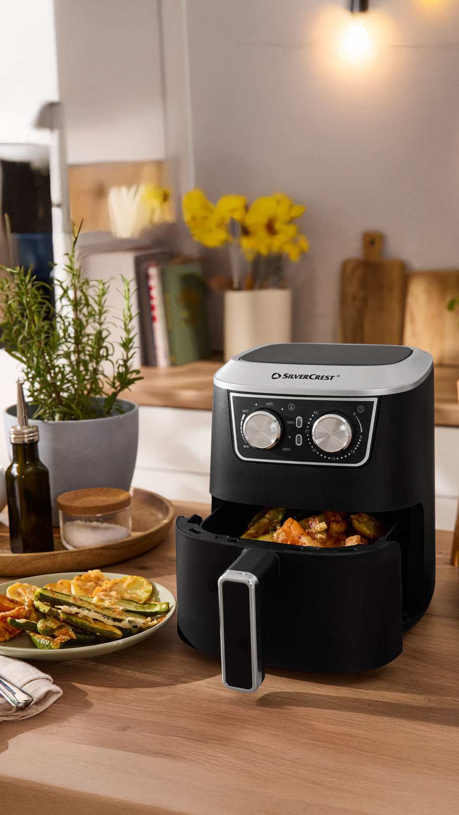 Friteuză cu aer cald neagră pe o masă de lemn, cu legume prăjite și ierburi aromatice.