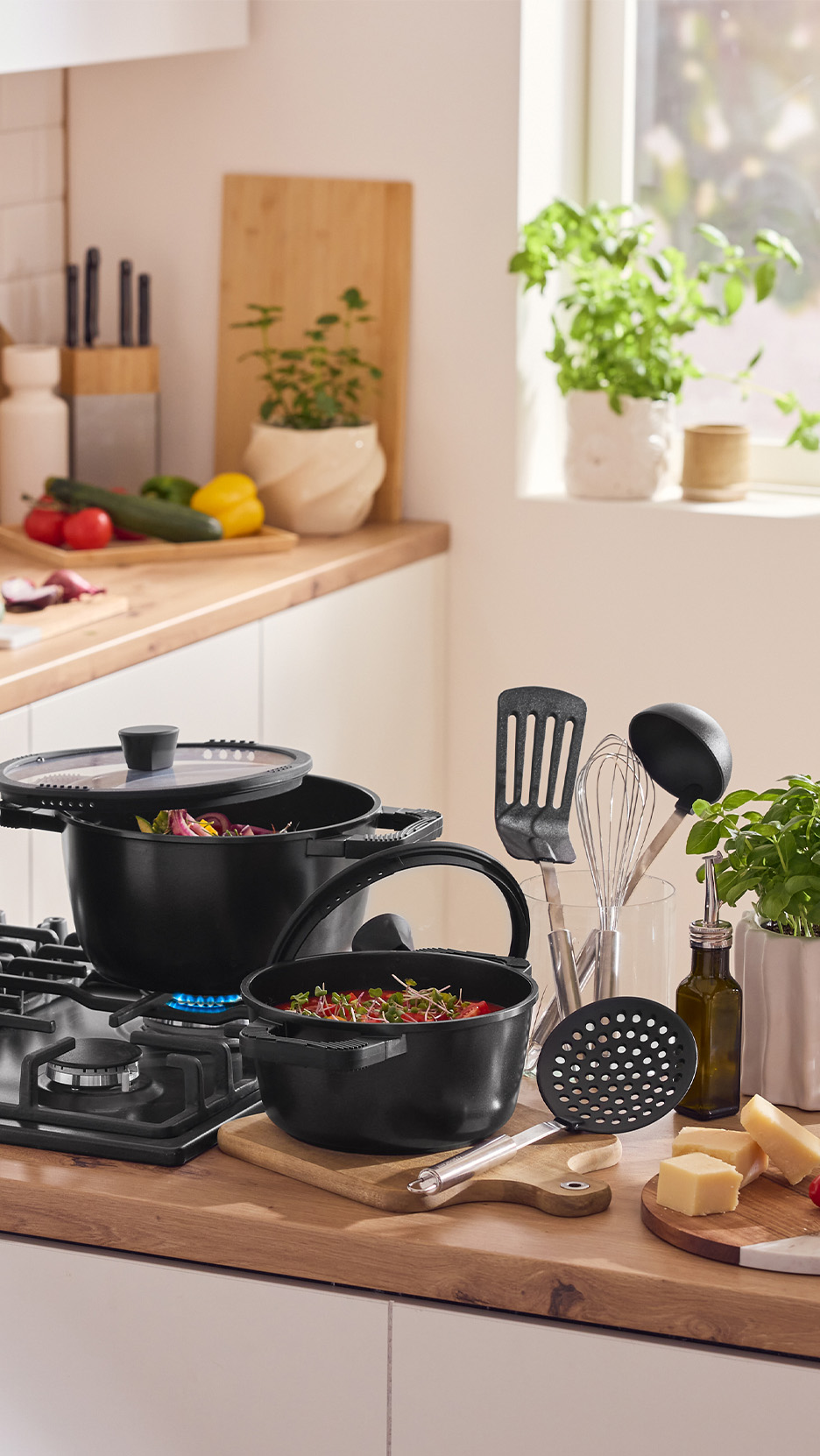 Oale și tigăi negre pe aragaz, cu ustensile de bucătărie și ingrediente proaspete.